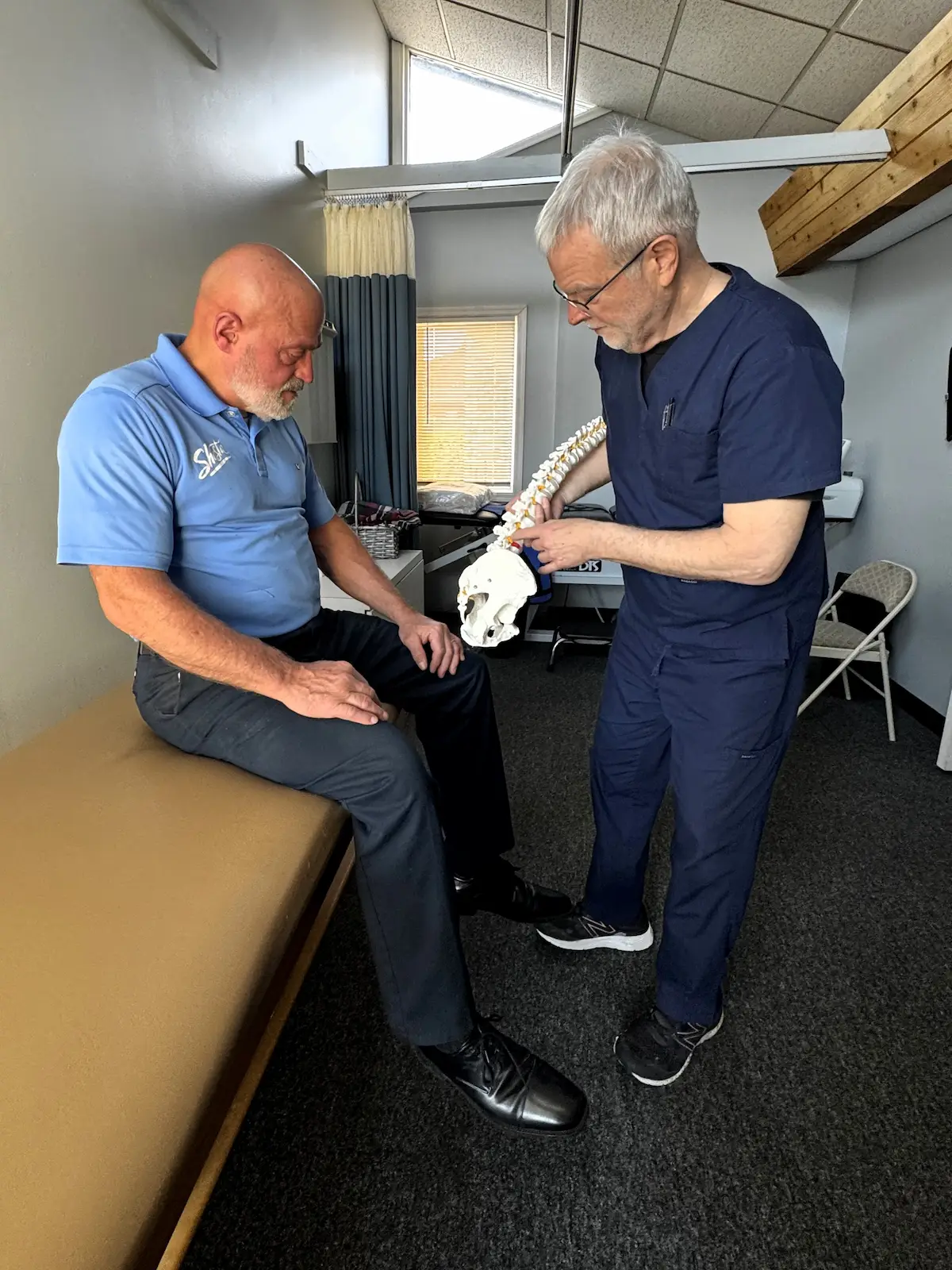 A healthcare professional in blue scrubs points to a spine model while explaining to a patient seated on an examination table.