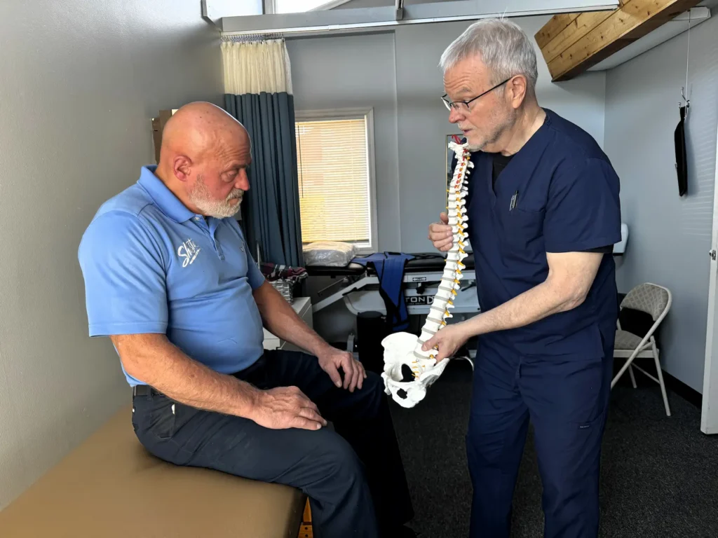 A healthcare professional in blue scrubs explains a spine and pelvis model to a seated patient in a medical office.