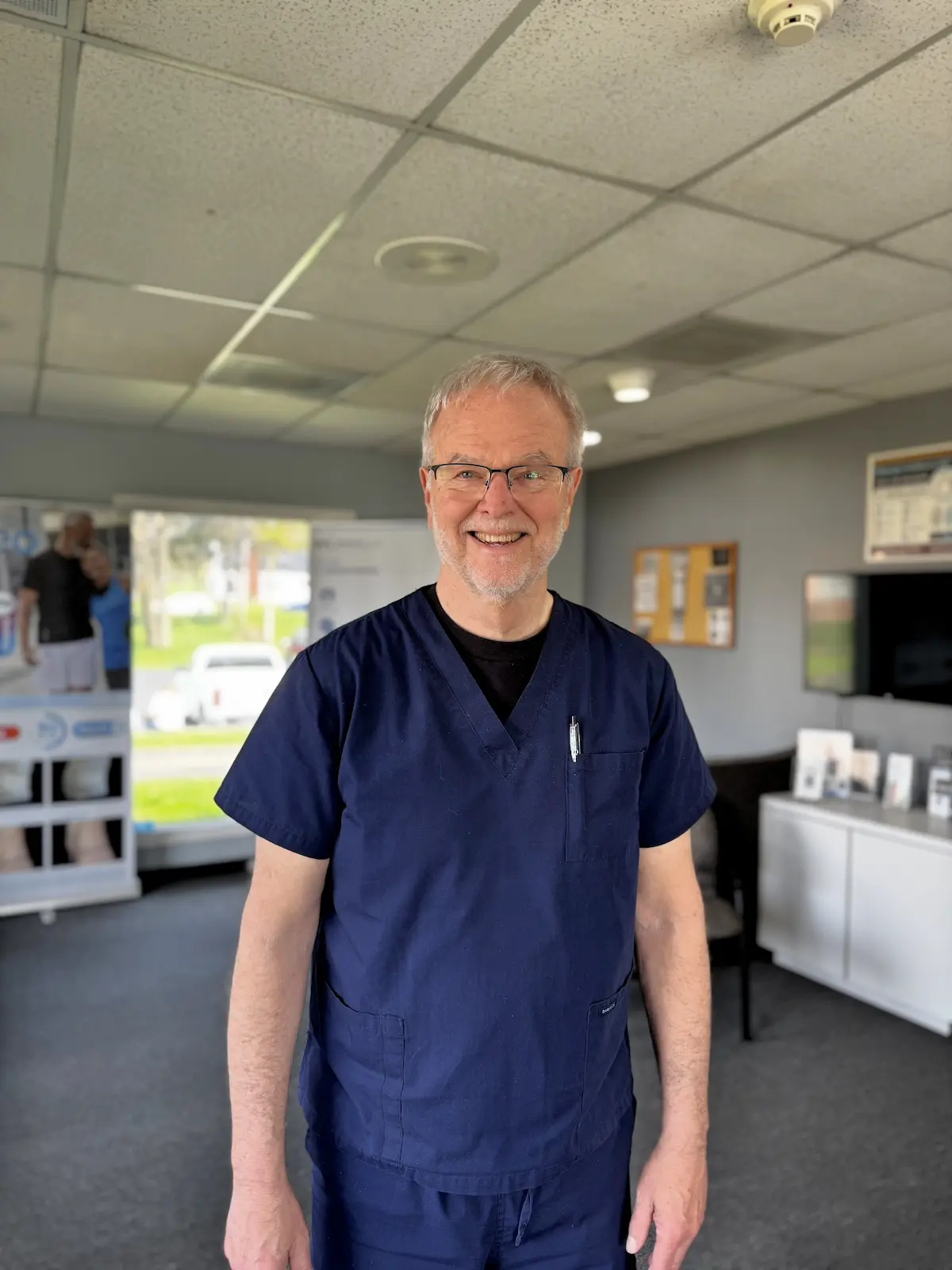 A smiling man with gray hair and glasses, wearing navy blue scrubs, stands in a brightly lit medical office.