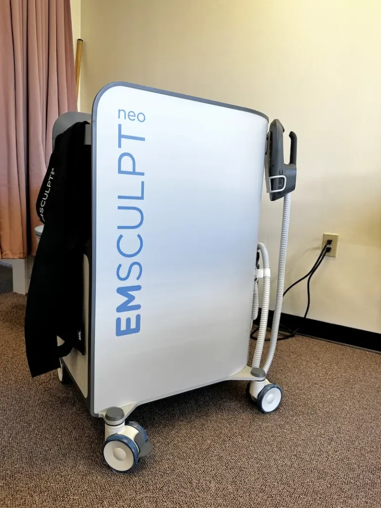 A white and gray Emsculpt Neo machine with blue lettering and an attached applicator, standing on a brown carpet.