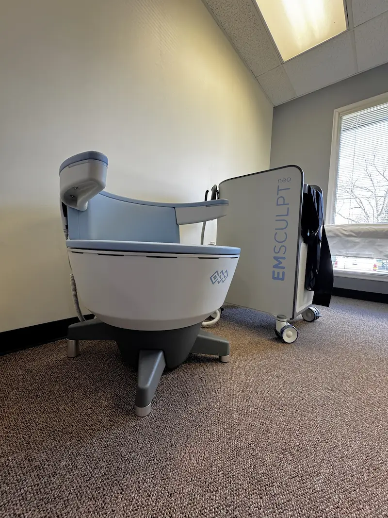 An EMSCULPT neo machine, white and light blue, stands on a brown carpet in a room with light walls and a window.