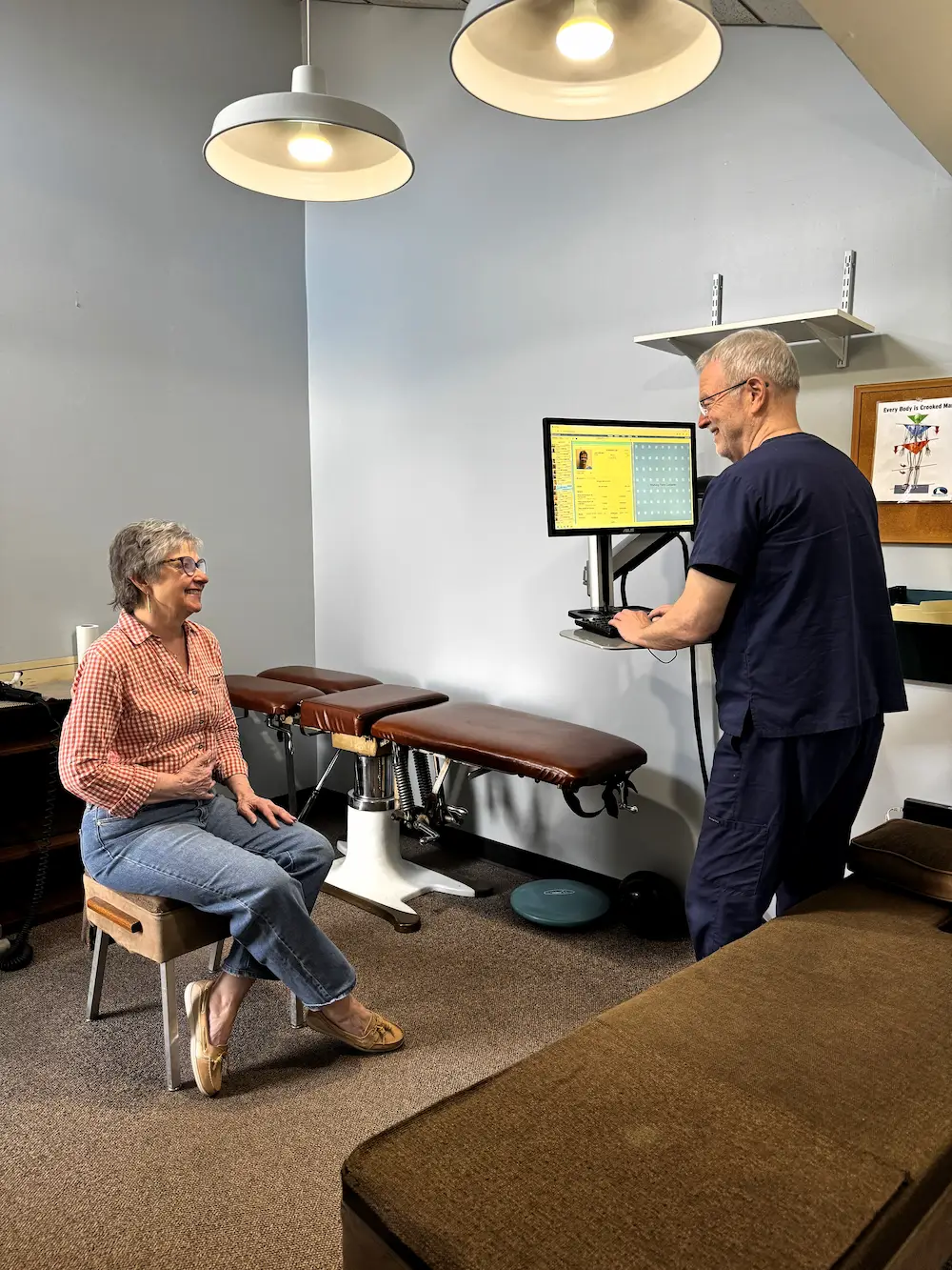 An older woman sits on a stool smiling at a male practitioner standing at a computer in a medical examination room.