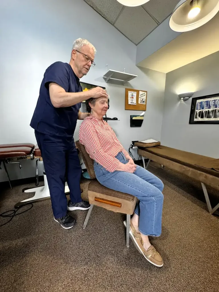 A male chiropractor adjusts the head and neck of a seated female patient in a clinic.