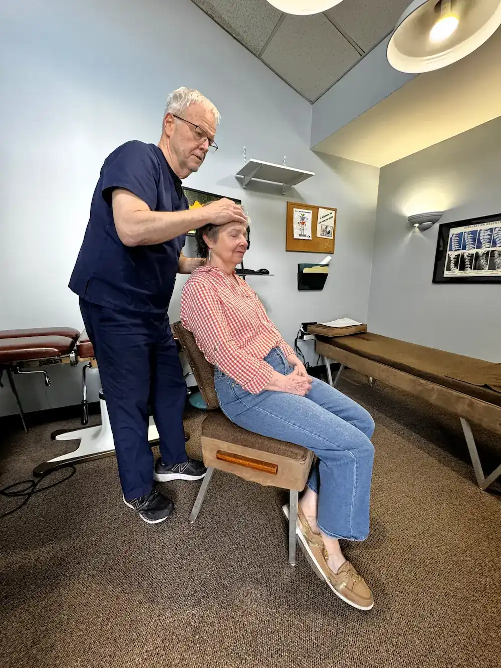 A male chiropractor adjusts the head and neck of a seated female patient in a clinic.