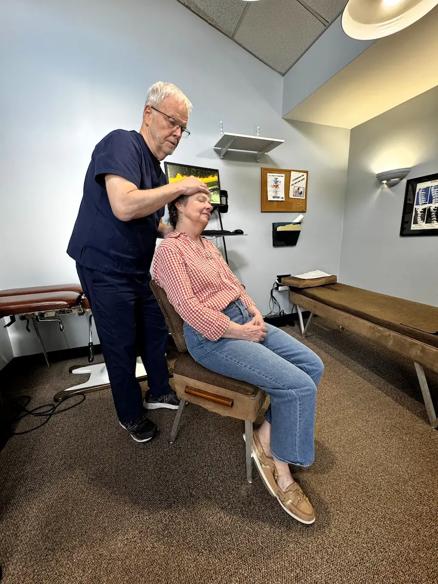 A male chiropractor with glasses stands behind a female patient seated in a chair, gently placing his hands on her forehead.