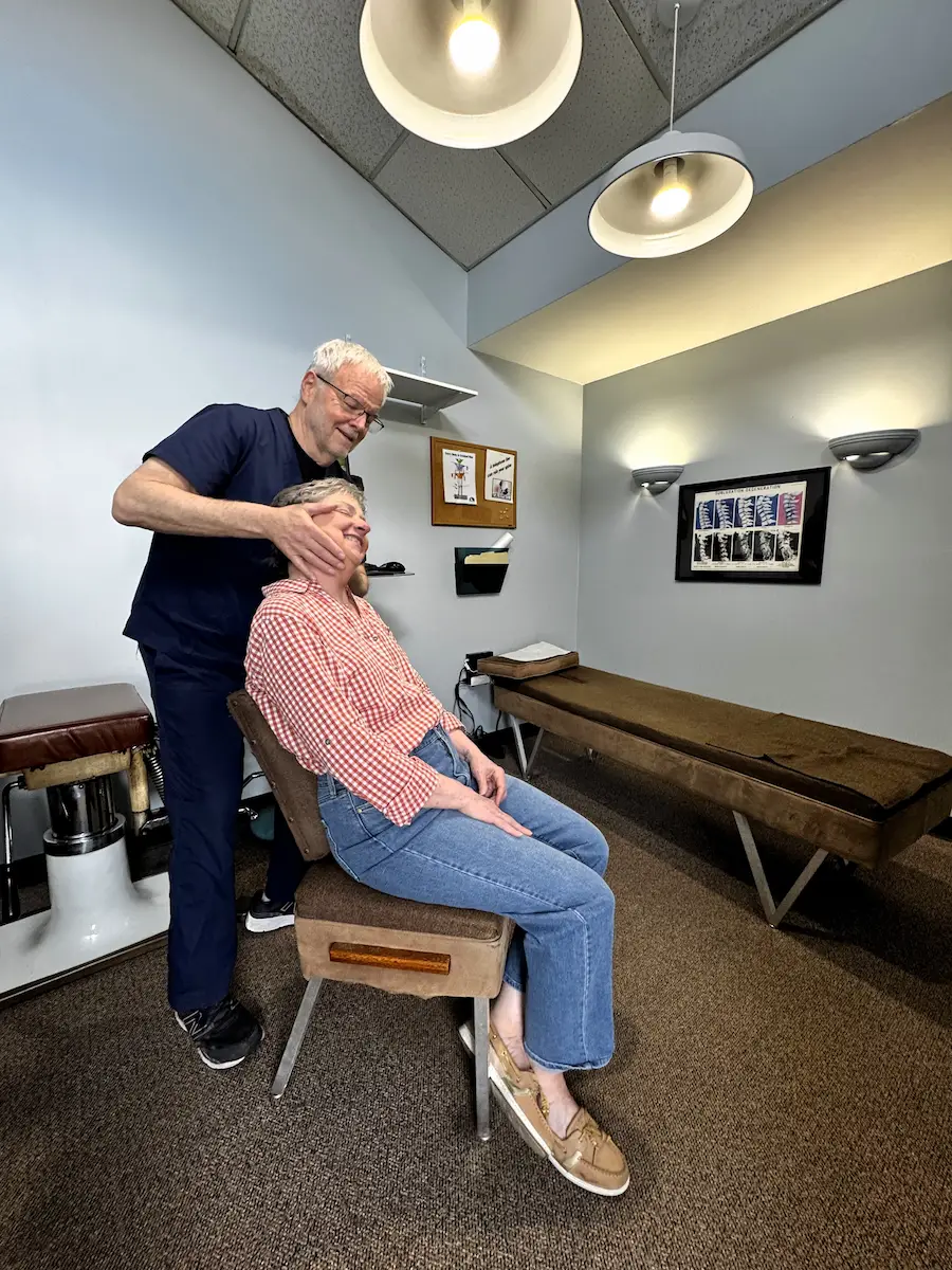 A male chiropractor in blue scrubs adjusts the neck of a smiling female patient seated in a chair, in a chiropractic office.