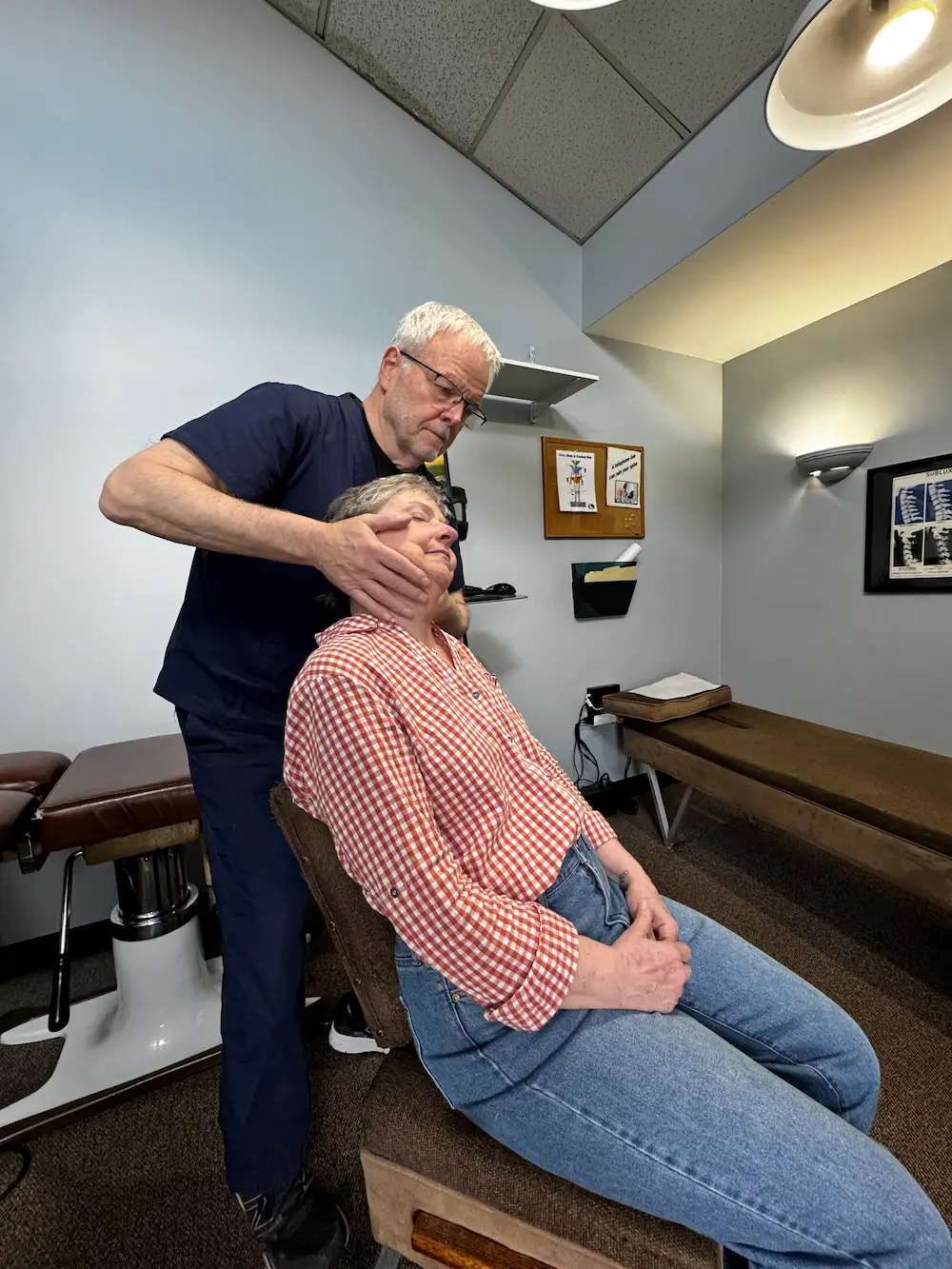 An older man in scrubs adjusts an older woman's neck while she sits in a chair in a medical office.
