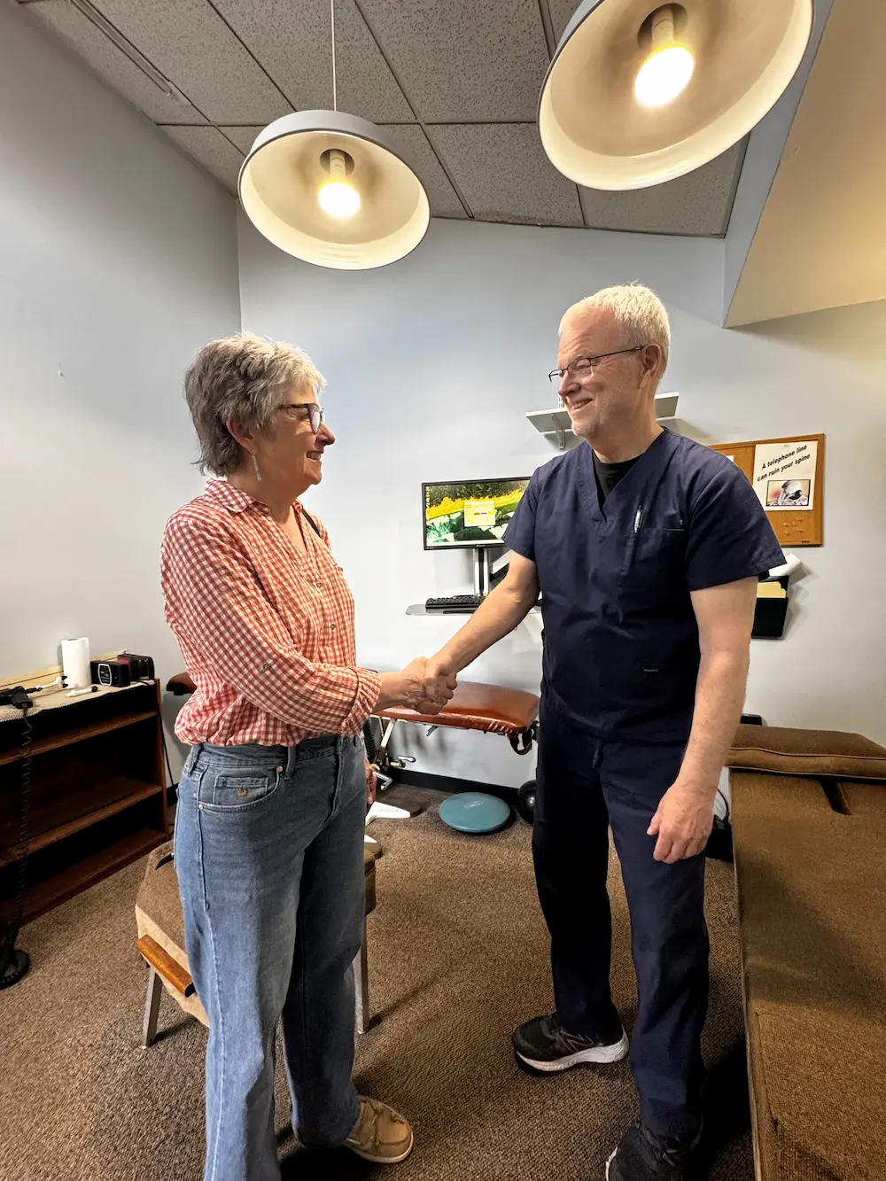 A woman in a checkered shirt and jeans shakes hands with a man in blue scrubs in a clinic office.