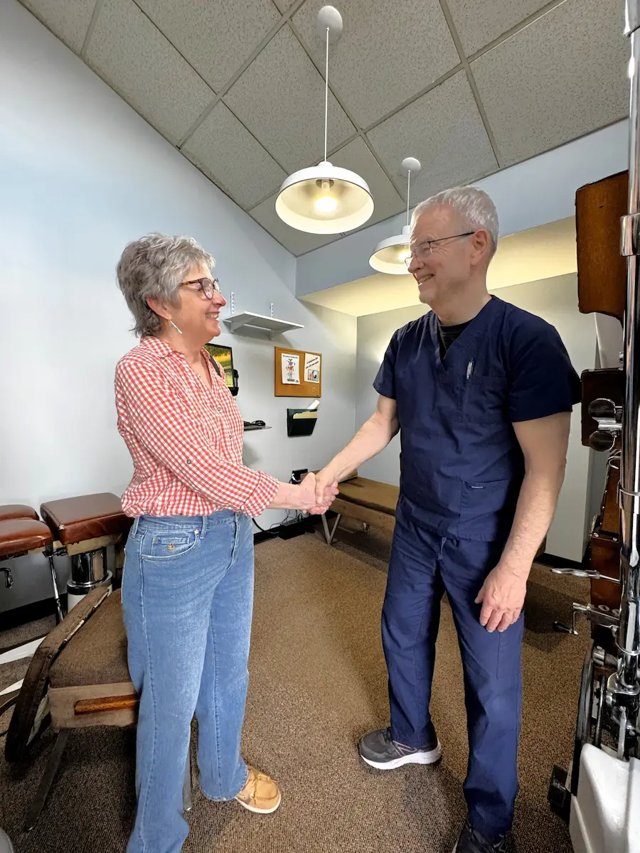 An older woman in a checkered shirt shakes hands and smiles with a man in blue scrubs in a medical office.