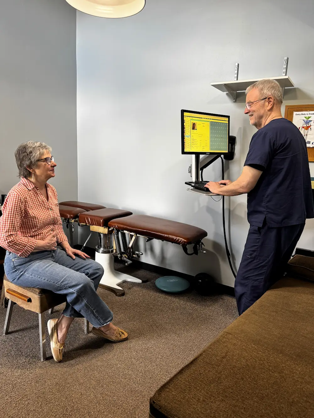 A doctor in blue scrubs stands at a wall-mounted computer, smiling at a woman seated on a stool in a chiropractic office.