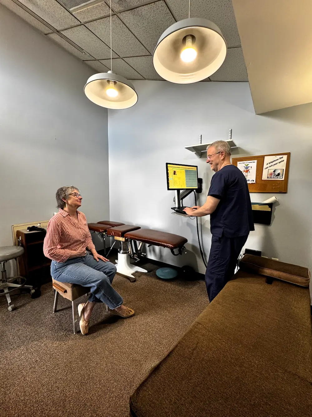 A woman in a plaid shirt sits on a stool, talking to a smiling man in blue scrubs who is looking at a computer screen in a chiropractic office.