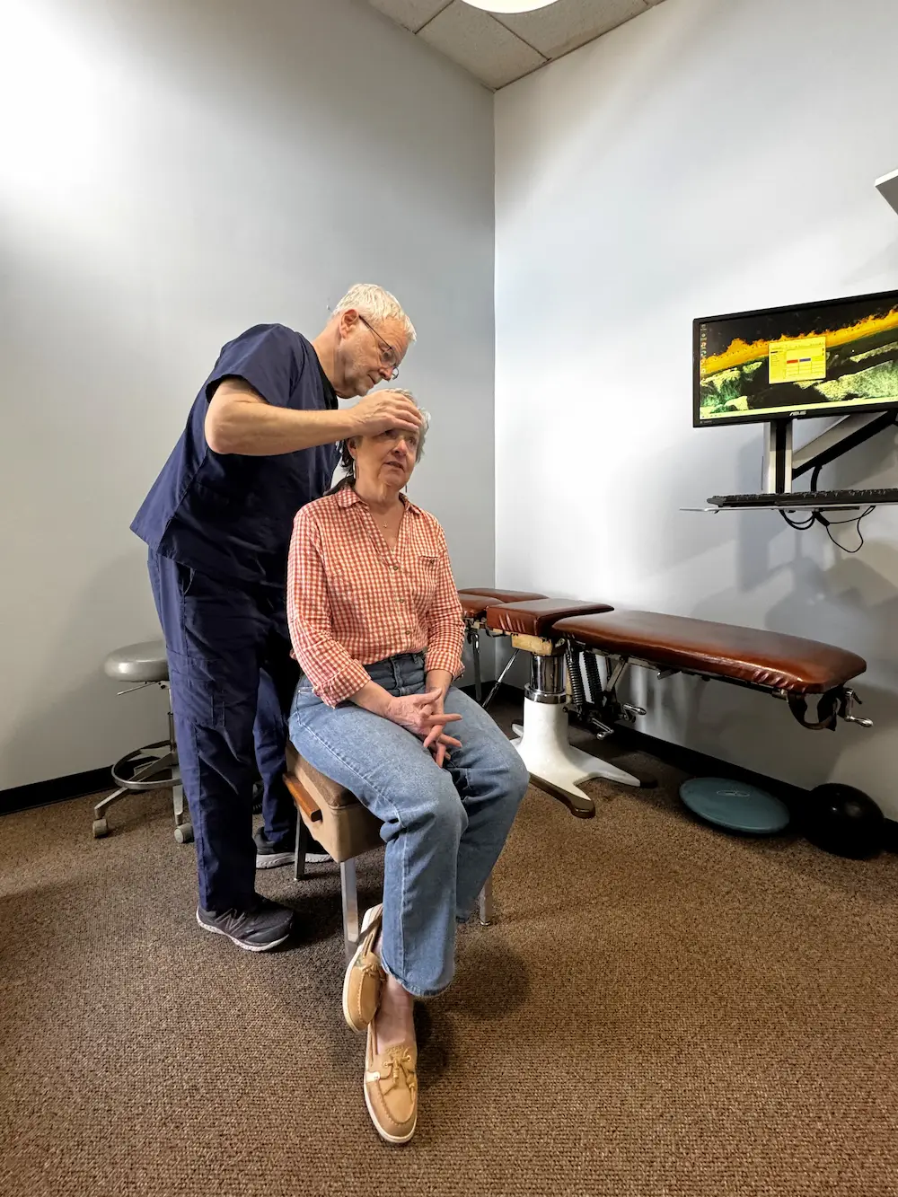 A male practitioner in blue scrubs examines a seated female patient's head in a clinic.