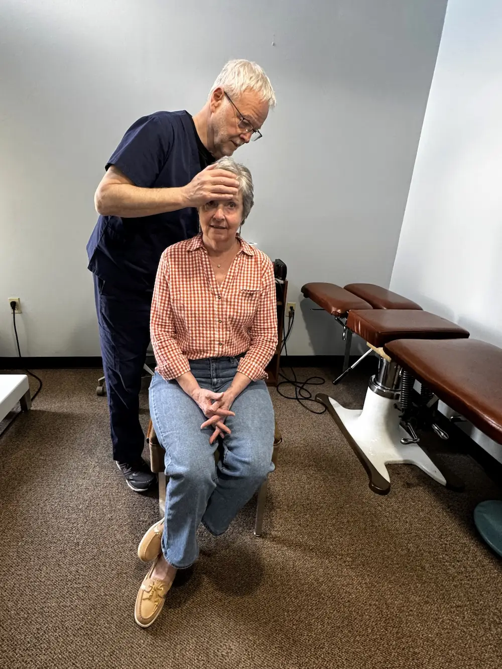 A male practitioner with gray hair and glasses stands behind a seated older woman, placing his hands on her head. She wears a checkered shirt and jeans.