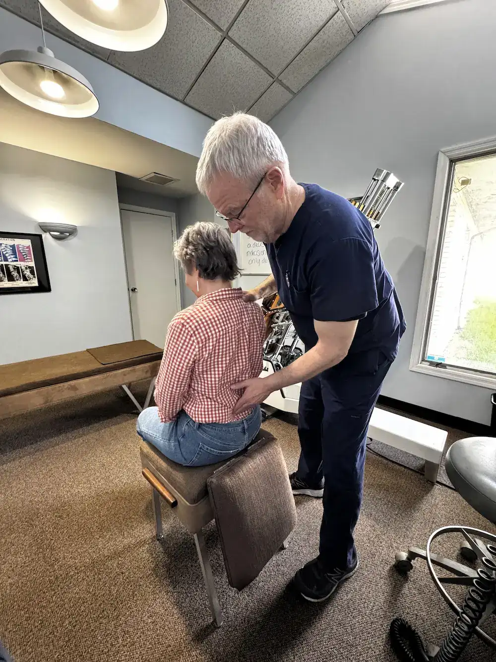 A male chiropractor in blue scrubs adjusts the lower back of a female patient seated on a stool in a medical office.