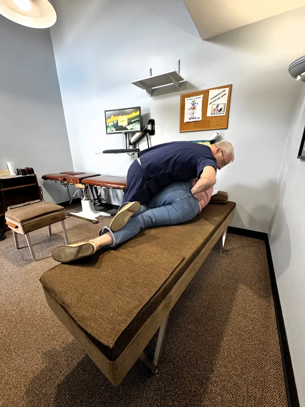 A chiropractor in blue scrubs adjusts a patient lying on their side on an examination table.