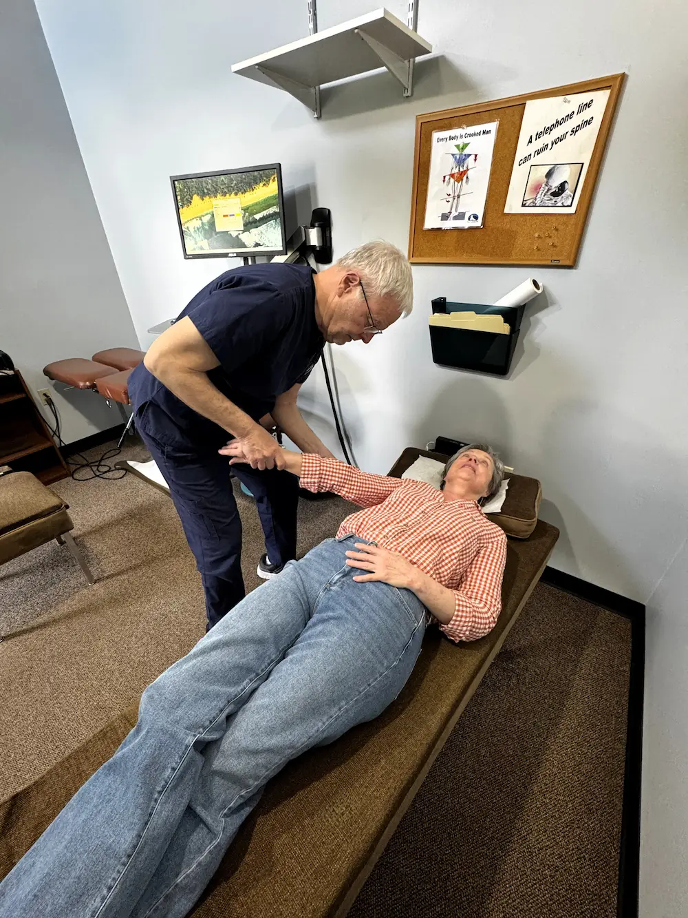 A male chiropractor adjusts a female patient's arm as she lies on a brown examination table in a clinic.