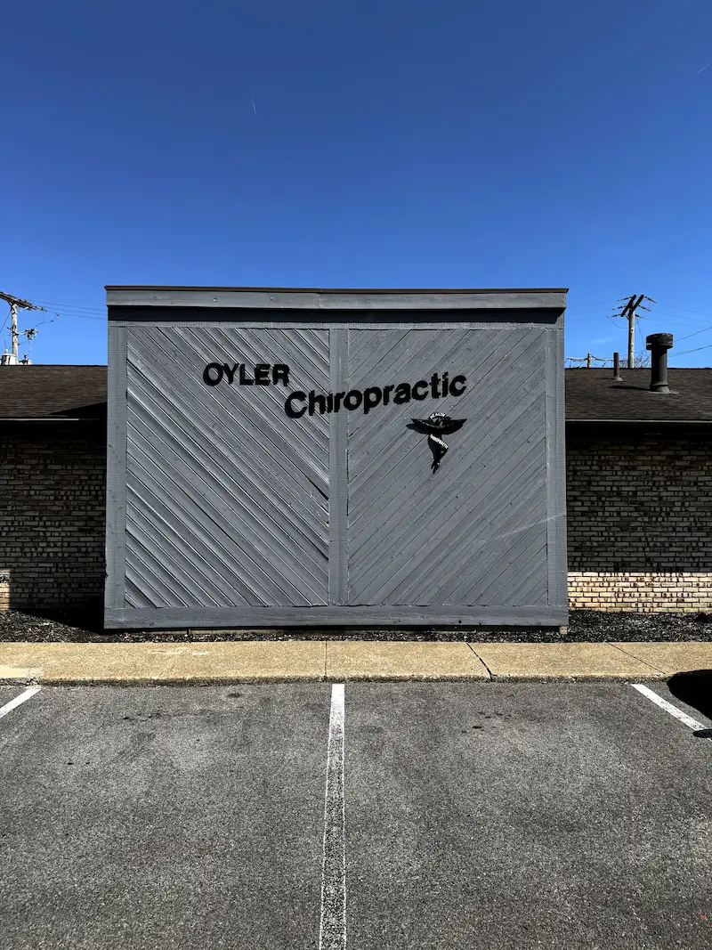 Exterior sign for OYLER Chiropractic on a gray wooden facade with diagonal paneling. The sign includes a caduceus symbol. A parking lot is in the foreground, and a clear blue sky is above.
