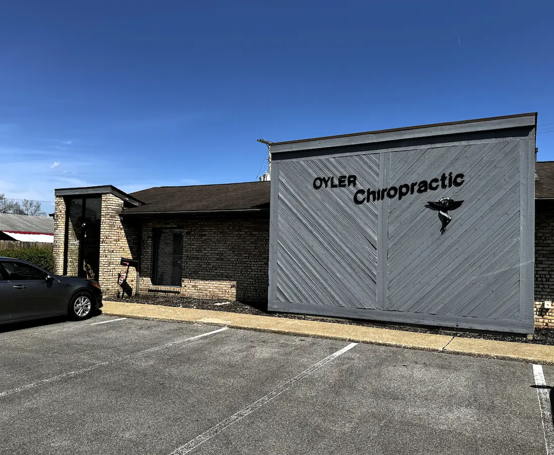 Exterior view of OYLER Chiropractic building with a gray wooden facade, brick walls, and a dark gray car in the parking lot.