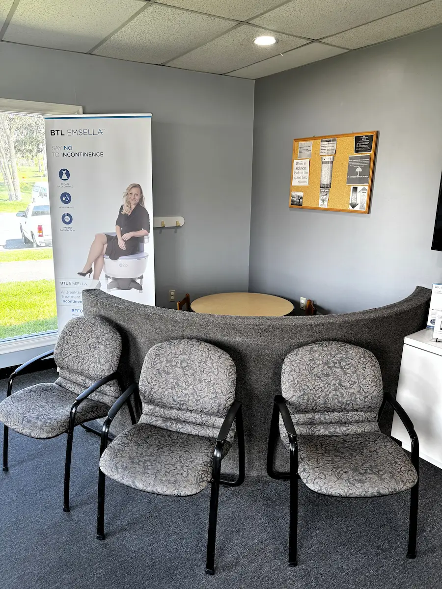 A waiting room with three grey chairs, a BTL Emsella banner, and a cork board on a grey wall.