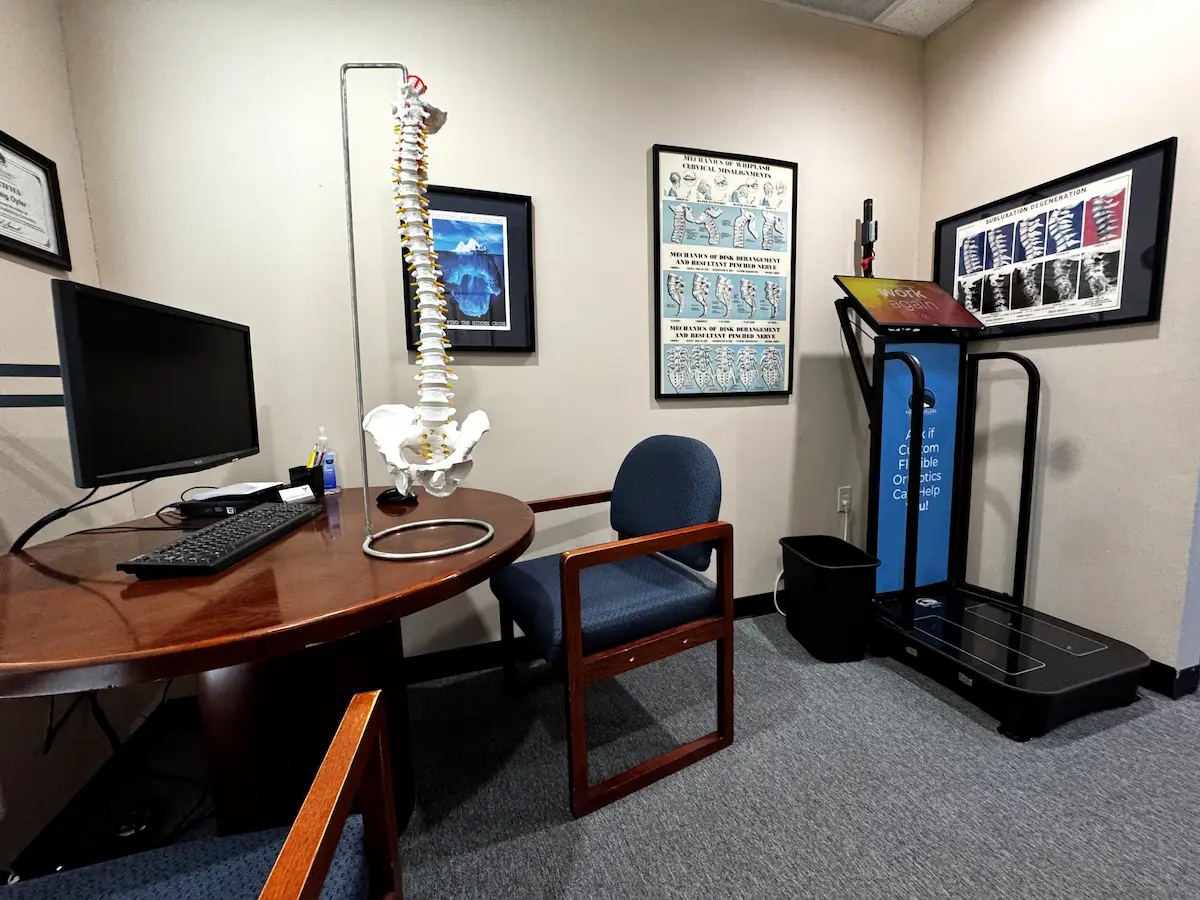 An office with a desk, computer, human spine model, anatomical charts, and a foot scanner machine, suggesting a chiropractic or orthotics clinic.