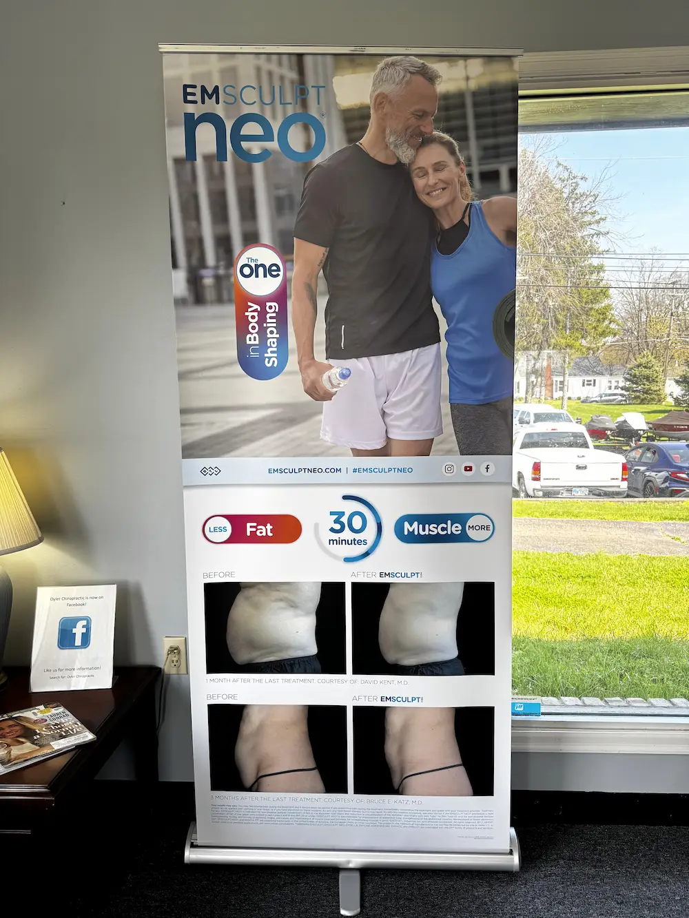 A vertical Emsculpt Neo banner showing a fit couple, text about fat reduction and muscle gain, and before-and-after photos of body shaping results. It stands in an indoor setting next to a table with a lamp and a window looking out onto a sunny street with vehicles and houses.