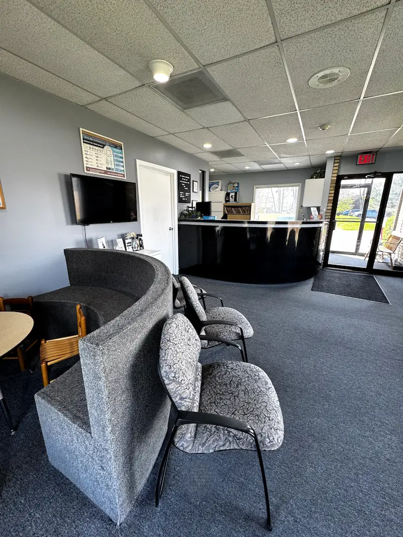 A waiting room with a curved gray bench, patterned chairs, a black reception desk, and a wall-mounted TV.