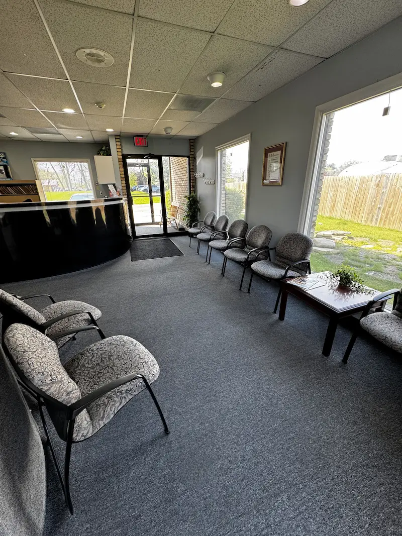 A waiting room with a black reception desk, gray carpet, and two rows of patterned chairs. Large windows look out to a grassy area and fence.