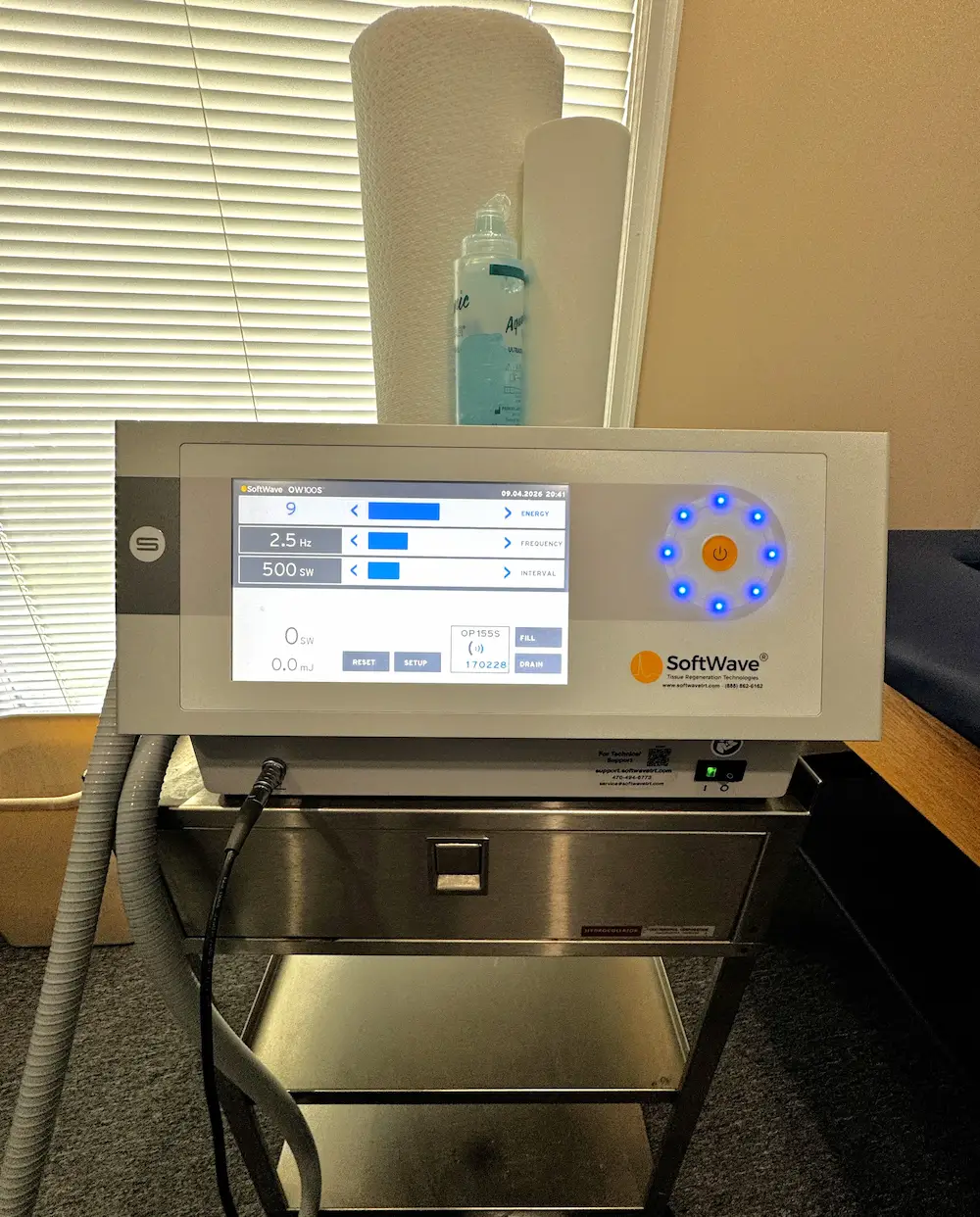 A SoftWave OW100S medical device on a stainless steel cart, with its digital screen displaying settings and a circular blue LED control panel. A bottle of ultrasound gel and paper towels are visible behind it.