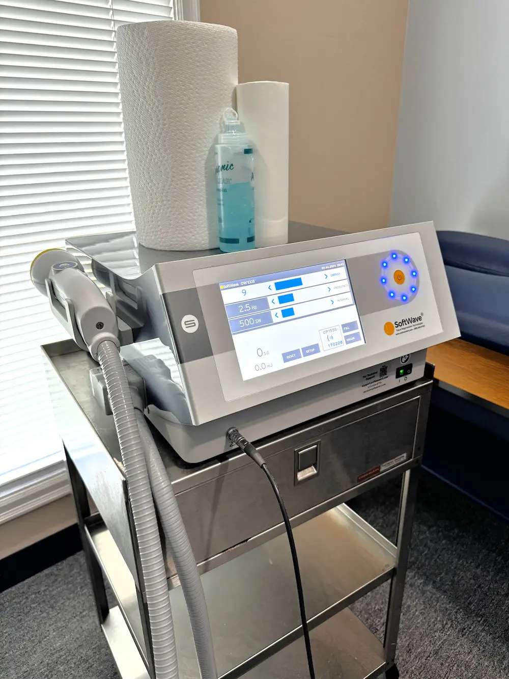 A SoftWave medical device on a stainless steel cart with a handheld applicator, digital display, paper towels, and blue gel.