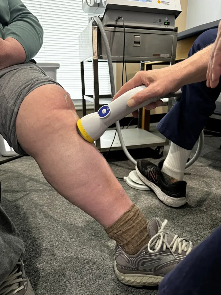 A handheld medical device applying treatment to a person's knee, with gel visible on the skin. Medical equipment is in the background.