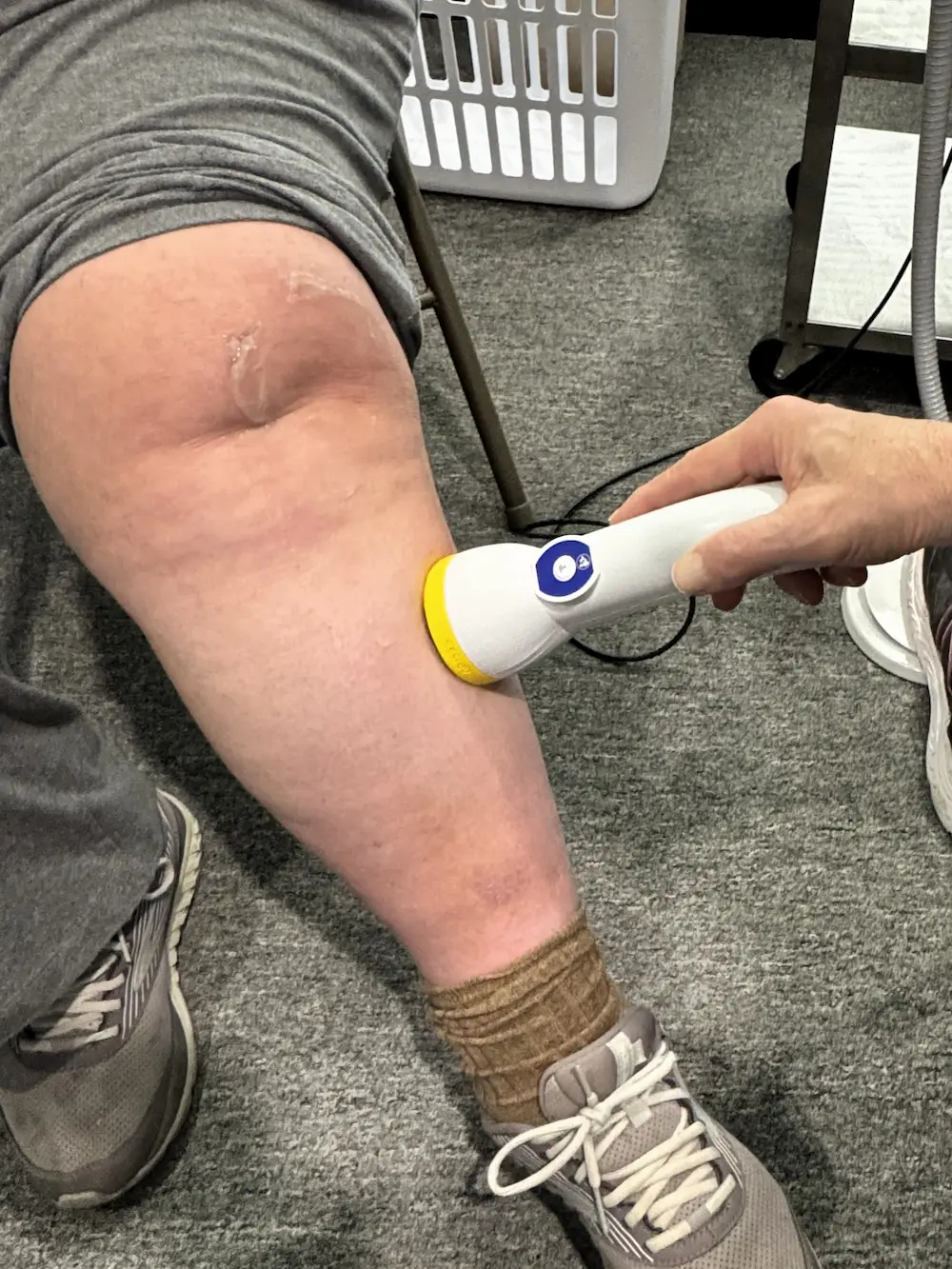A close-up of a person's leg with a handheld white and yellow medical device being pressed against the skin just above the knee. Clear gel is visible on the knee.
