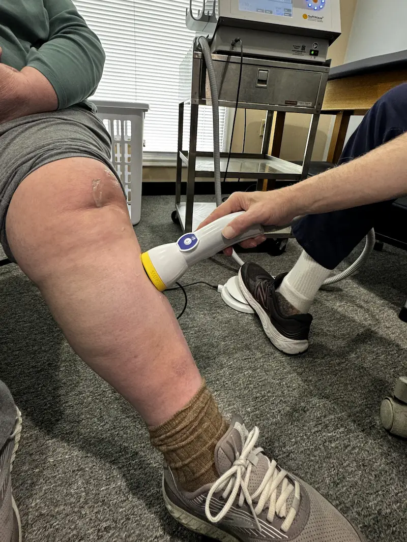 A person's leg receiving treatment with a white and yellow SoftWave therapy device on the lower leg, just below the knee. Clear gel is on the knee.