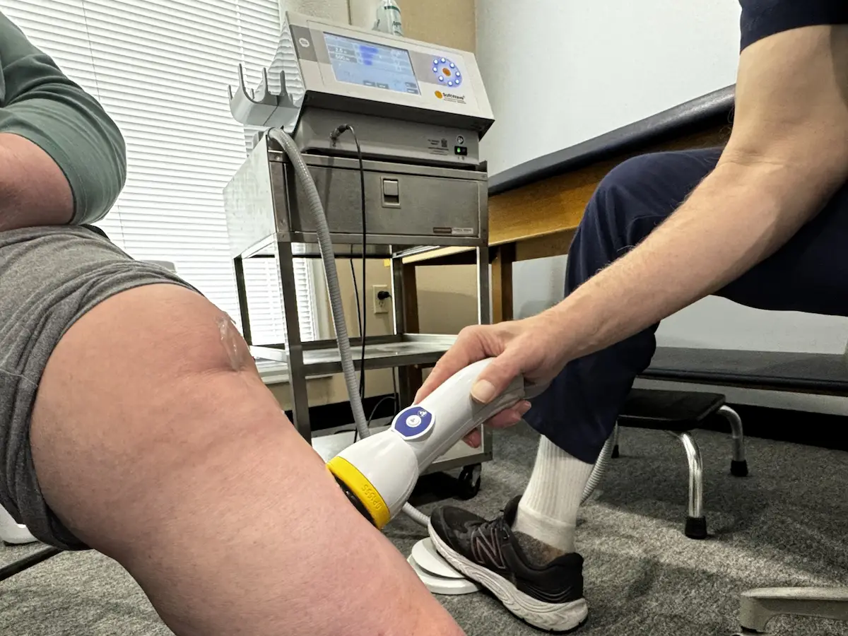 A medical professional applies a handheld therapy device to a patient's knee, which has clear gel on it. A medical machine is visible on a cart in the background.