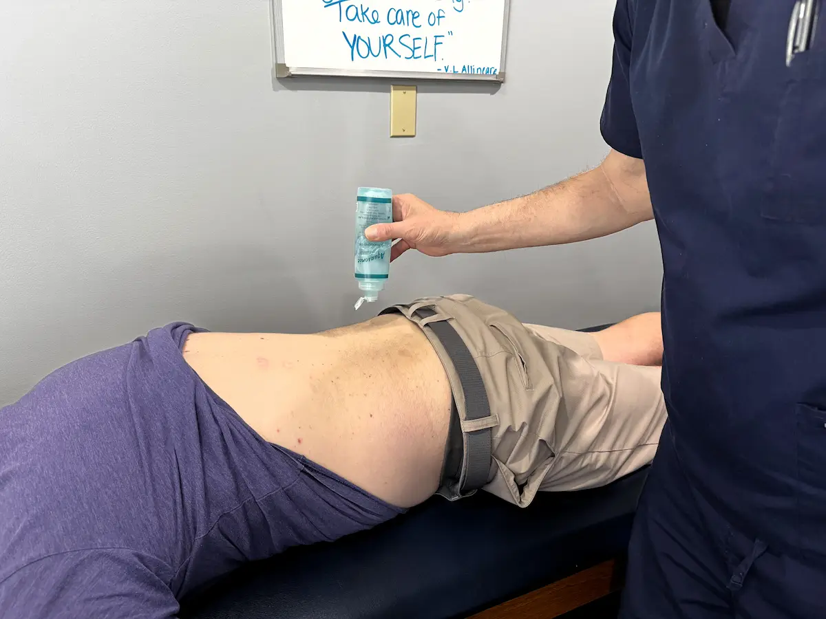 A medical professional applies ultrasound gel to a patient's lower back on an examination table.