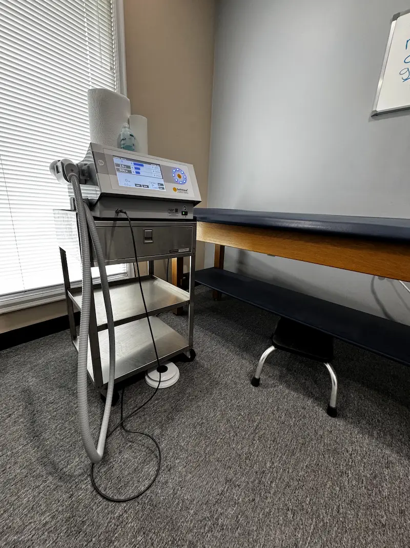 A grey and silver medical device on a stainless steel cart with a digital screen, hoses, and cables, positioned in a room with a dark blue examination table, a window with blinds, and a whiteboard.
