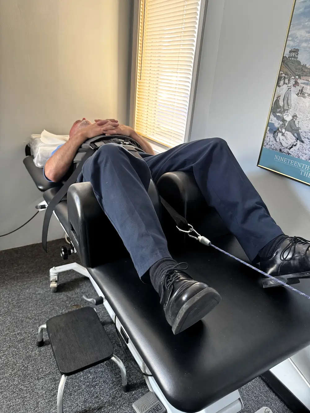 A person lies on their back on a black medical table, secured by straps and a harness, with a rope attached to their feet area.