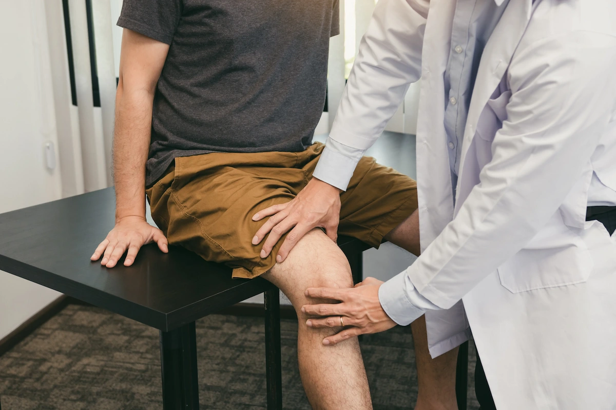 A doctor in a white lab coat examines a patient's knee, with both hands on the knee.