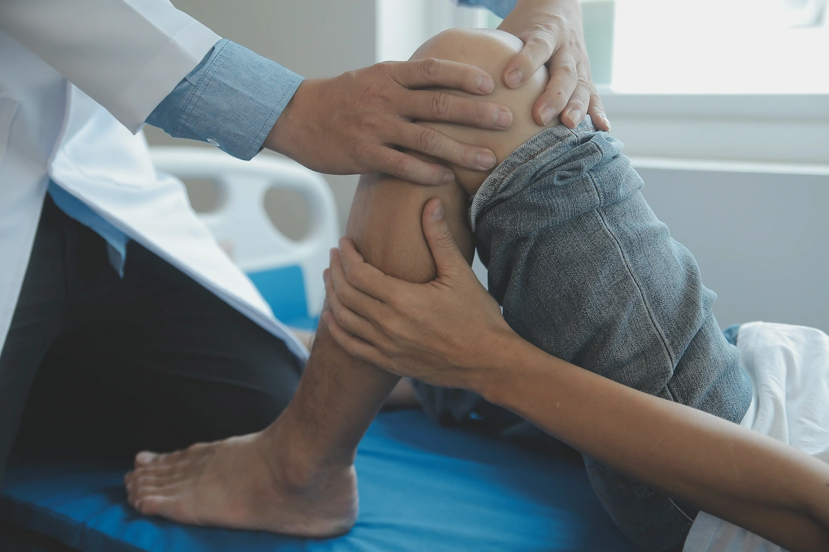 Close-up of a doctor's hands examining a patient's bent knee on an examination bed.